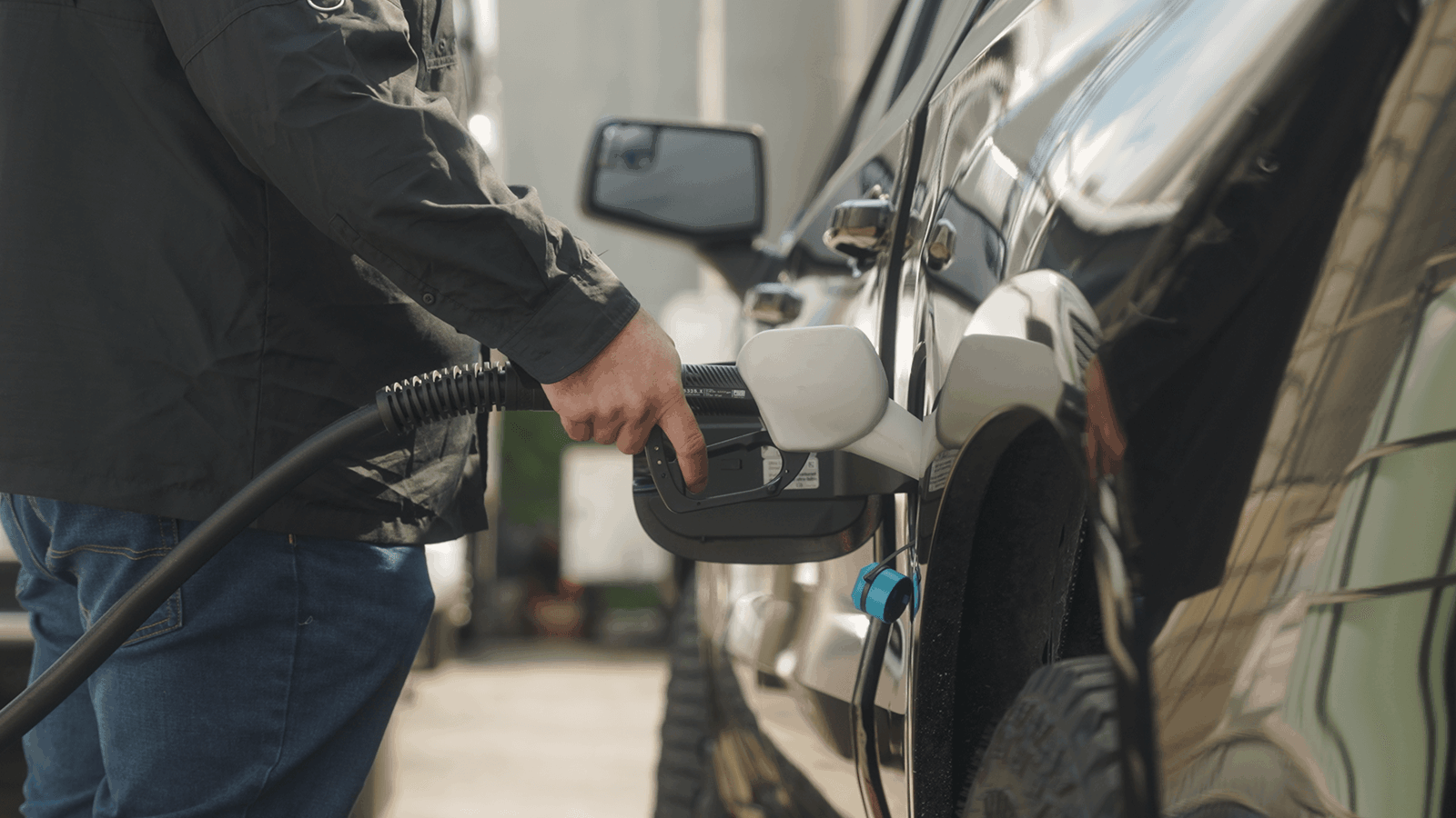 En person laddar ett svart elfordon utomhus genom att ansluta en kontakt liknande AdBlue pumphandtag med flödesmätare till bilens port, vilket betonar effektiv energiöverföring.
