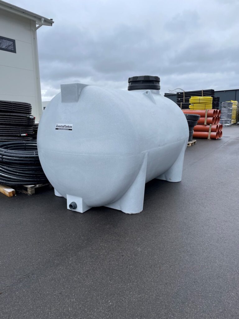 En ÄlvestadTanken Vattentank LBO 7500-10400 Liter Livsmedelsgodkänd, en stor vit horisontell vattentank i plast med en cirkulär topplucka, står på en asfalterad yta utomhus med kablar och material i bakgrunden under en molnig himmel.