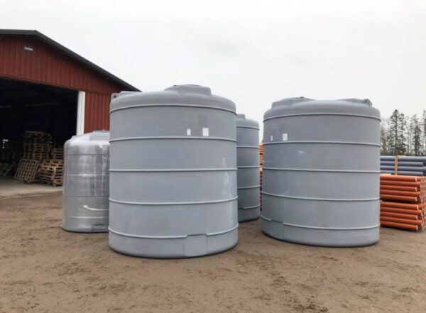 Flera stora Vattentank 10 kubik - uthyres-enheter står utomhus på en grusplan nära en röd metallbyggnad, med orange rör och ytterligare tankar synliga i bakgrunden under en mulen himmel.