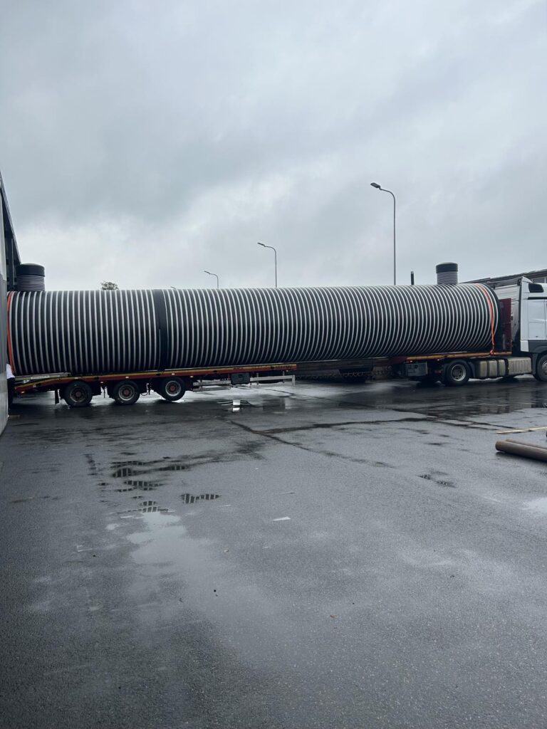 Ett långt, cylindriskt, svartvitrandigt industrirör lastas på ett lastbilsflak i närheten av tre Vattentankar 75 000 liter i Västerbotten en våt, mulen dag, med gatlyktor och byggnader synliga i bakgrunden.