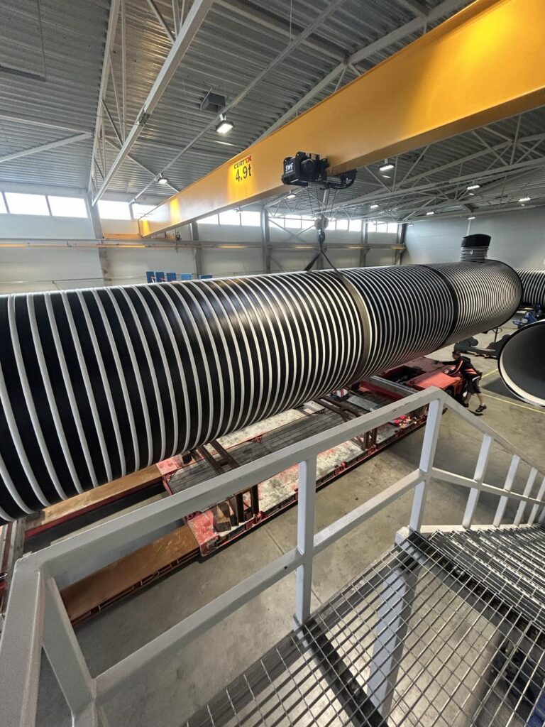 Ett stort cylindriskt objekt, en del av projektet 3 x Vattentankar 75 000 Liter - Västerbotten, med svarta och vita spiralränder, hänger i en kran inne i en industrilokal med metallräcken och högt i tak.