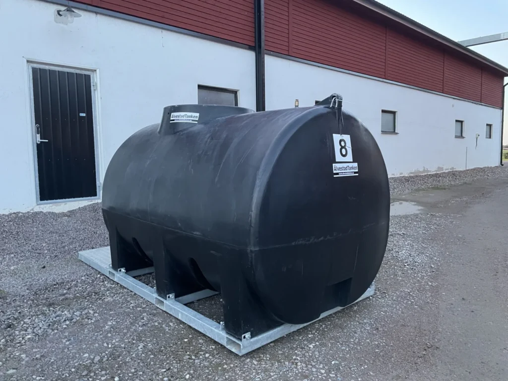 En Vattentank 5000 liter på ramverk är placerad på en betongplatta utanför en vit byggnad med svart dörr och röda lister, omgiven av grus.