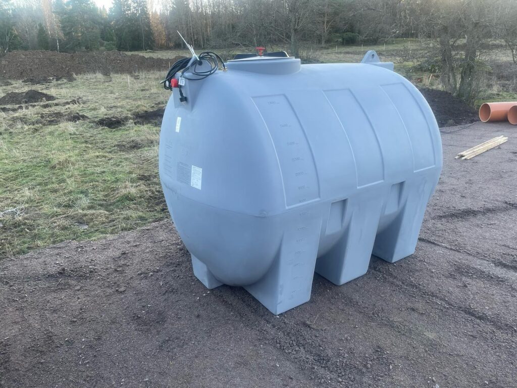 Vattentank 3000 liter med dränkbar pump & värmekabel är en stor grå cylindrisk plasttank med mätmarkeringar, placerad utomhus på jord och omgiven av gräs, träd och byggmaterial.