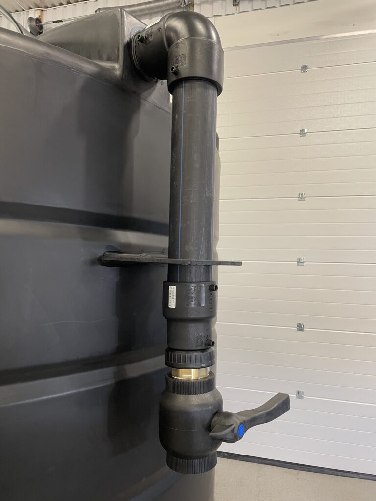 Närbild på en Sugledning med kulventil - en svart vattentank med ett stort svart PVC-rör och en avstängningsventil med blått handtag - i en industri- eller lagermiljö.
