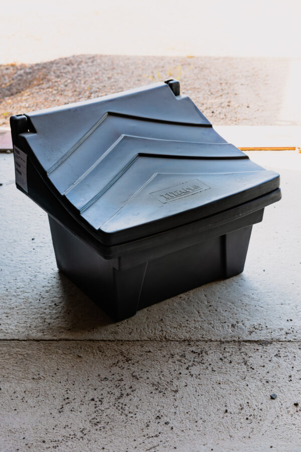 En stor svart Sandlåda förvaringslåda 50 liter med ett texturerat, vinklat lock vilar på ett betonggolv i ett garage, upplyst av solljus som strömmar in genom en öppen dörr.