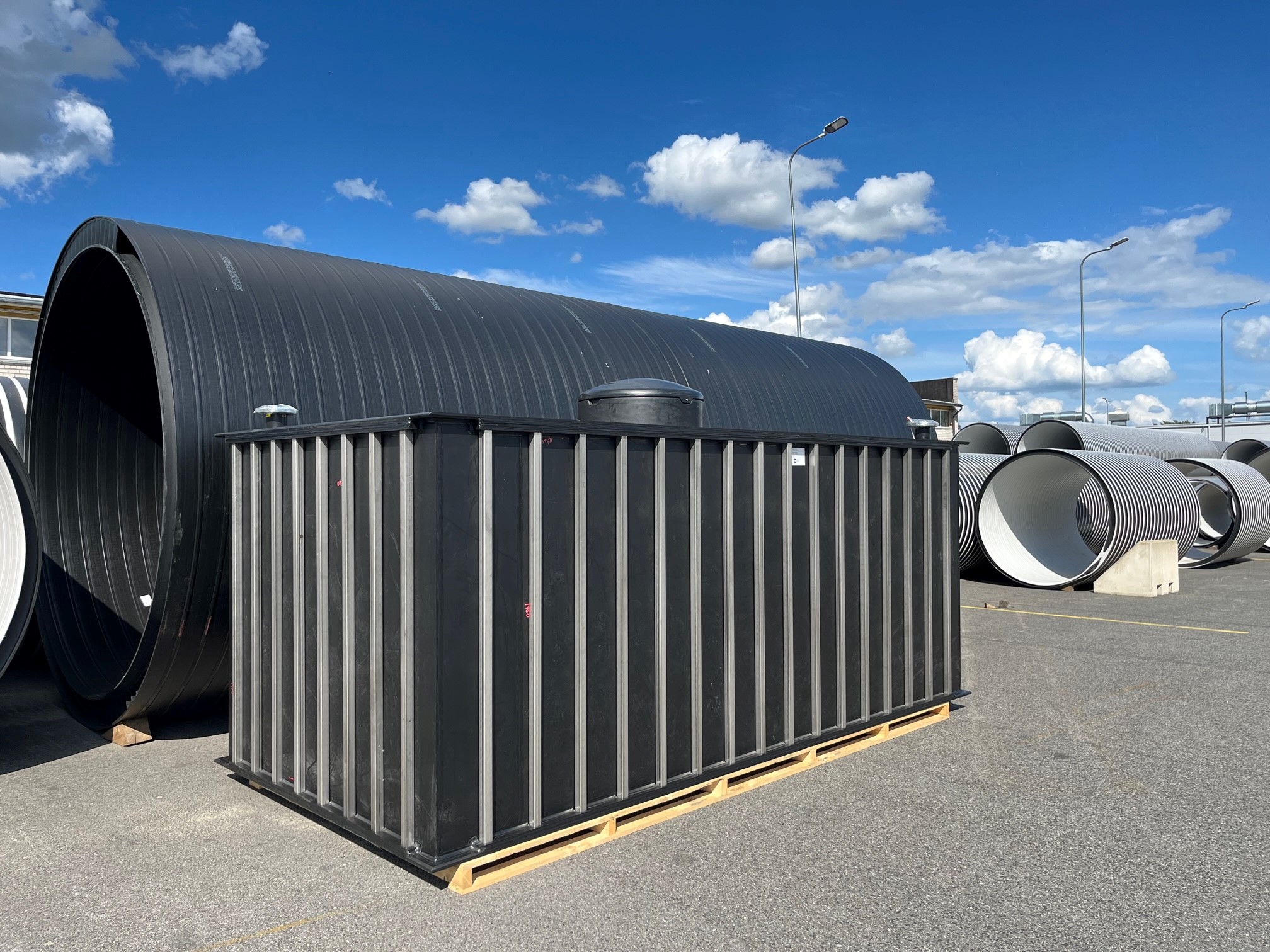 Stora svarta plasttankar och rör, bland annat en måttanpassad 15 000 liters regnvattentank, står utställda utomhus på en asfalterad yta under en ljusblå himmel med spridda moln. En rektangulär tank är placerad på en träpall i förgrunden.