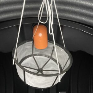 En nätkorg med metallram hänger i vita snören under en orange ÄlvestadTanken regnvattentank 10.000 liter inne i en mörk, rundad struktur.