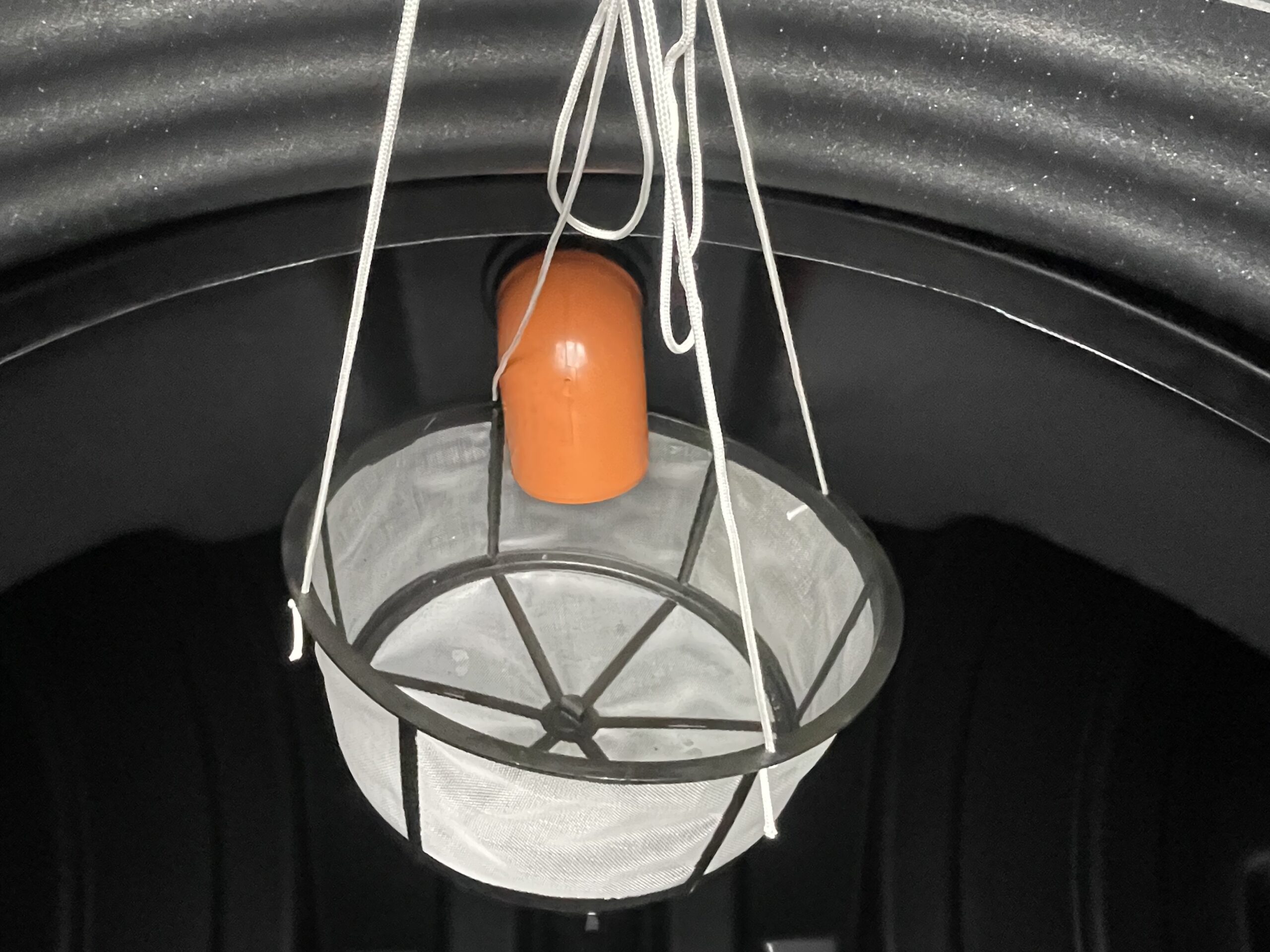 En nätkorg med metallram hänger i vita snören under en orange ÄlvestadTanken regnvattentank 10.000 liter inne i en mörk, rundad struktur.