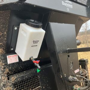 En närbild visar en jordbruksmaskin med ÄlvestadTanken Renvattentank 14 liter vit tank monterad, med synliga varningsetiketter och rörledningar i en lantlig utomhusmiljö.