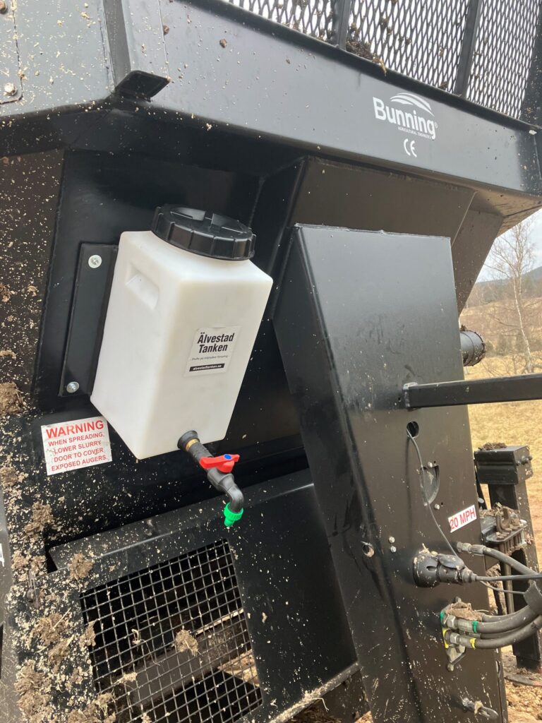 En närbild visar en jordbruksmaskin med ÄlvestadTanken Renvattentank 14 liter vit tank monterad, med synliga varningsetiketter och rörledningar i en lantlig utomhusmiljö.