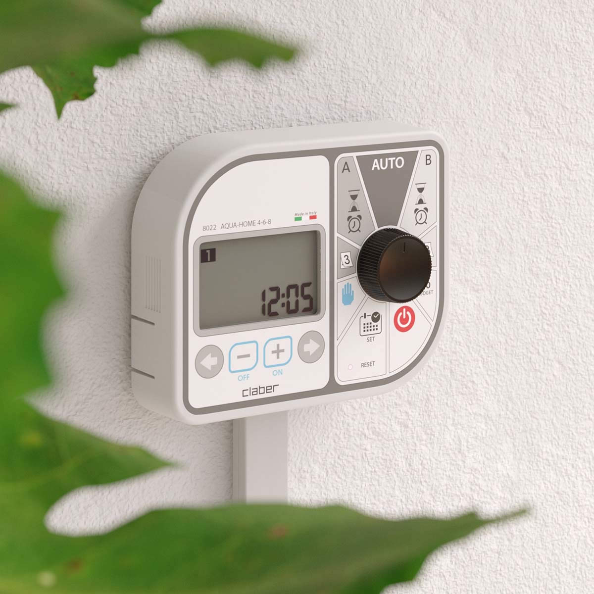 Claber vattentimer "AQUA-HOME 4-6-8" är monterad på en vit vägg och visar "12:05" på sin display. De olika knapparna och den stora svarta urtavlan är synliga, och i den vänstra förgrunden syns delvis en grön bladväxt.