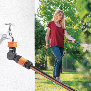 En kvinna i röd skjorta och jeans vattnar gräsmattan med en trädgårdsslang ansluten till en utomhuskran med Claber ledad krankoppling 1"-3/4". I bakgrunden skymtar gröna träd och solljus.
