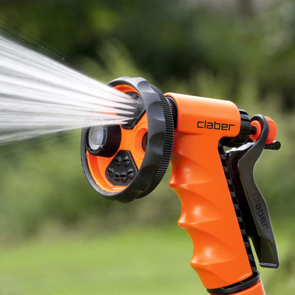 Närbild av en orange Claber "ERGO-GARDEN" spraypistol som avger en kraftig vattenstråle, mot en suddig grön bakgrund.