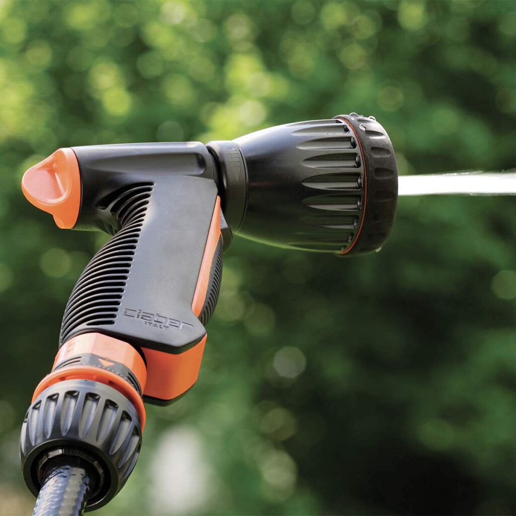 Närbild av Claber "MULTI-JET" strålpistol i svart och orange som sprutar vatten, med en suddig bakgrund av gröna bladverk.