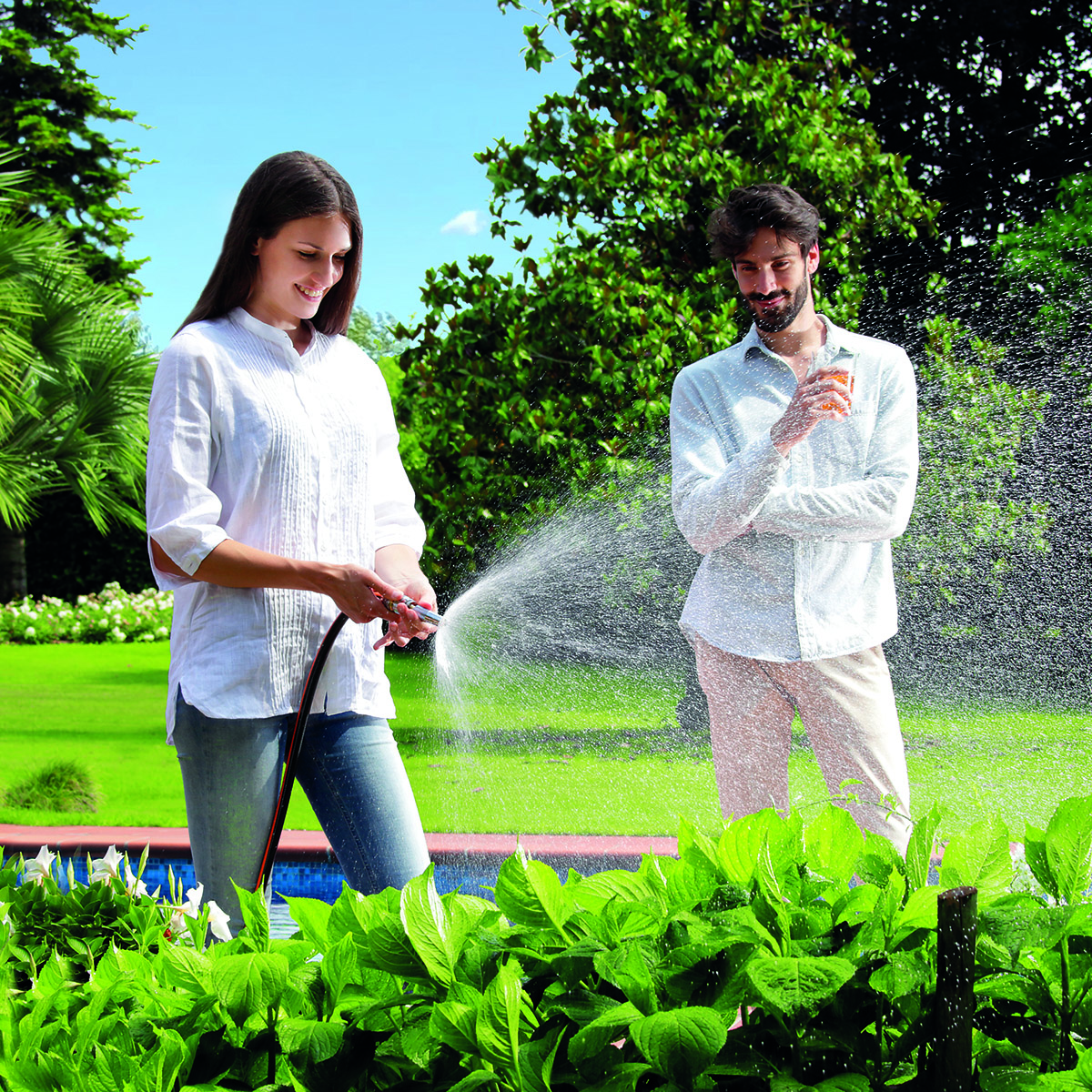 En kvinna använder Clabers strålpistol "Metal-Jet" för att vattna växter i en solig och grönskande trädgård, medan en man i närheten ler och tar en drink bland gröna träd och buskar.