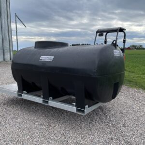 En svart Transporttank vatten 2000L ink ramverk med kupolformad topp står på en metallram över grus nära gröna fält och en hög silo under moln. Bakom tanken syns en del av en traktor eller ett nyttofordon.