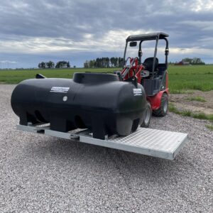 En liten röd lastare transporterar en Transporttank vatten 1200L bläckramverk på en metallplattform utomhus, med grus, gröna fält och en molnig himmel i bakgrunden.
