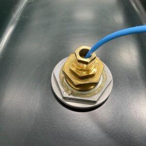 En blå kabel för Adapter R6 högnivågivare till spilloljetankar är fäst med en kabelförskruvning i mässing och monterad på en metallisk, mörkgrå yta.