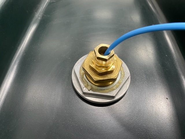 En blå kabel för Adapter R6 högnivågivare till spilloljetankar är fäst med en kabelförskruvning i mässing och monterad på en metallisk, mörkgrå yta.