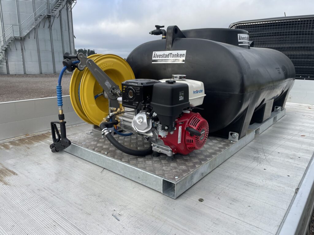 En stor svart bränsletank med slangvinda, pump och motor monterad på en metallplattform, placerad på baksidan av ett flakfordon. I bakgrunden syns en byggnad och en mulen himmel.