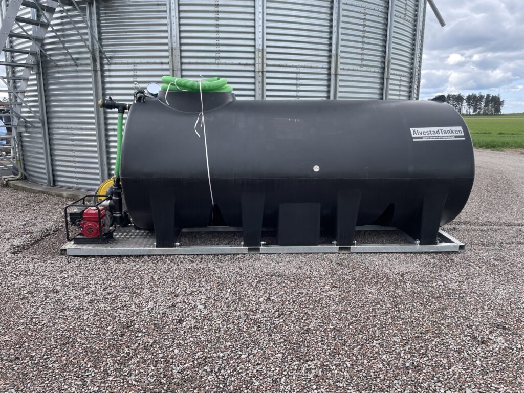 En stor svart cylindrisk tank märkt Avloppstanken är placerad på en grusyta bredvid en spannmålssilo i metall, med gröna slangar och en röd motor monterade. I bakgrunden syns öppen jordbruksmark och träd.