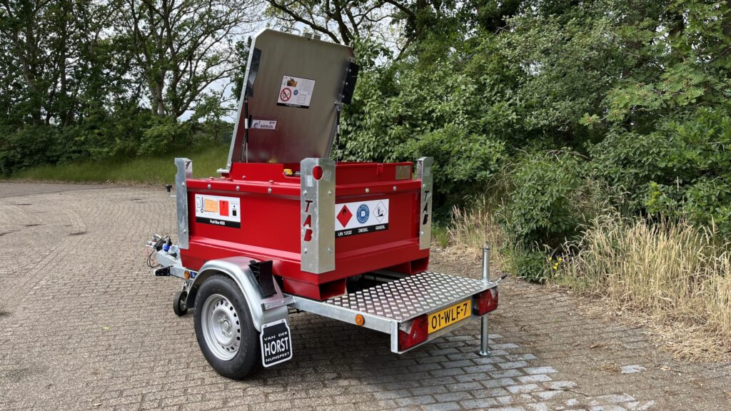 En röd bränsletrailer med öppet metallock står parkerad på en asfalterad yta nära gräs och träd. Släpet har varningsetiketter och en gul registreringsskylt på baksidan.