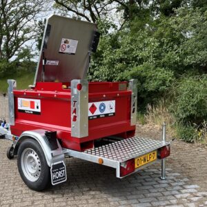 En röd bränsletrailer med öppet metallock står parkerad på en asfalterad yta nära gräs och träd. Släpet har varningsetiketter och en gul registreringsskylt på baksidan.