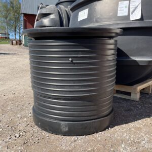 En Förhöjning till Aquabank Sluten Tank är installerad på en stor svart plasttank utomhus på grus, tillsammans med liknande tankar, med en lada och träd i bakgrunden under en blå himmel.