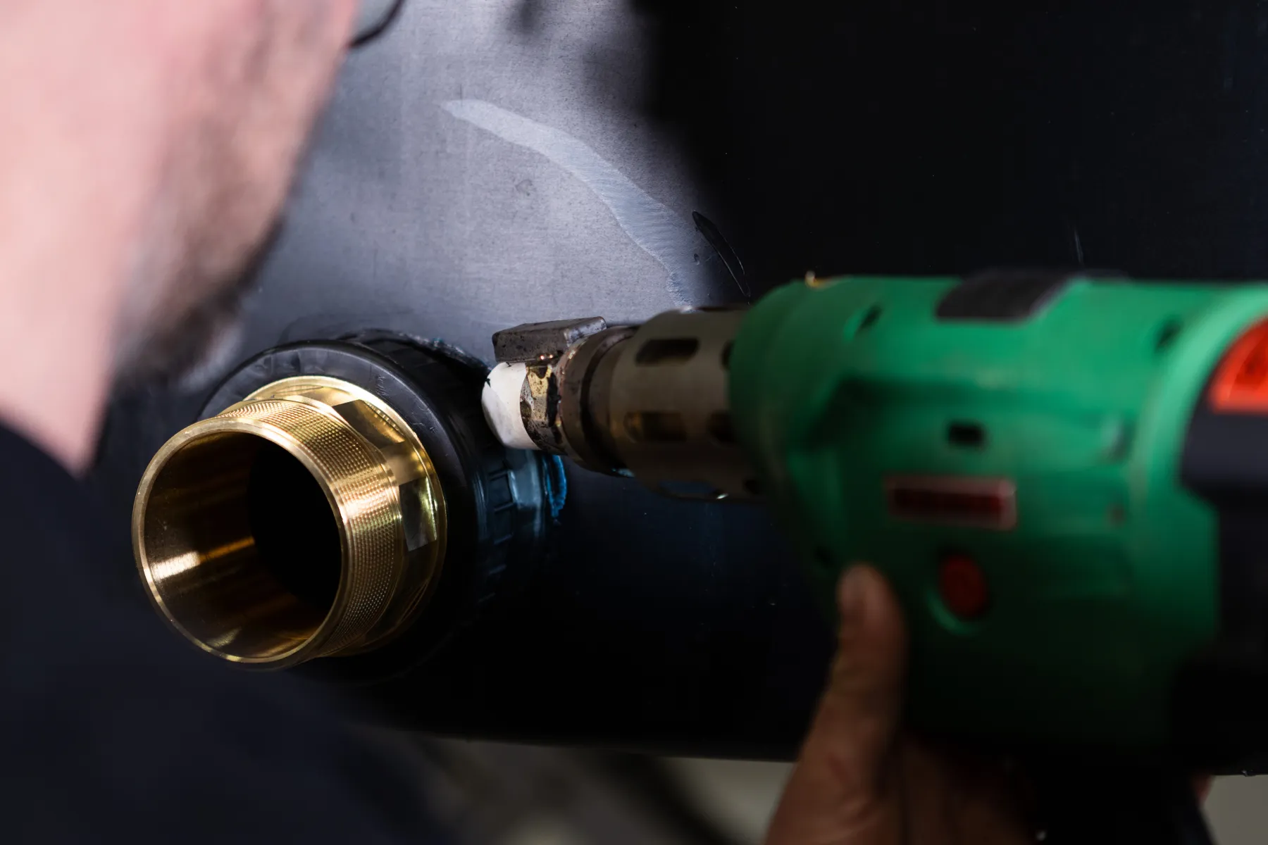 Kundspecifik tillverkning En person använder en grön värmepistol för att svetsa eller täta ett mässingsbeslag på en svart yta, möjligen en del av en VVS- eller industriinstallation.