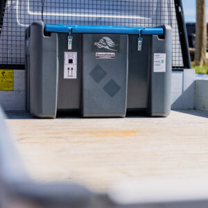 En TruckMaster KOMBI 400/40 Diesel/AdBlue förvaringscontainer med blått handtag och etiketter är säkrad på flaket till en lastbil, med nätskydd och träd i bakgrunden.