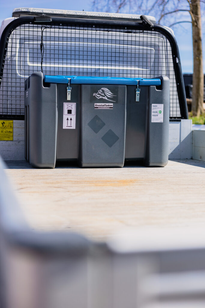 En TruckMaster KOMBI 400/40 Diesel/AdBlue förvaringscontainer med blått handtag och etiketter är säkrad på flaket till en lastbil, med nätskydd och träd i bakgrunden.