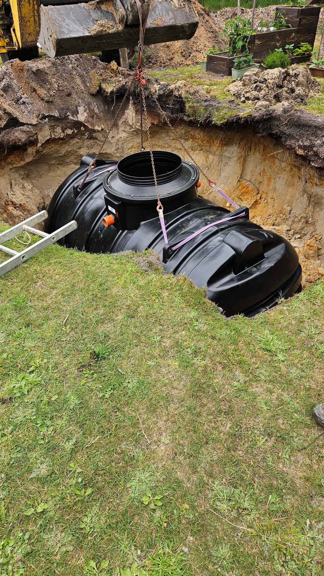 En ÄlvestadTanken regnvattentank på 10.000 liter sänks ned med spännband i ett djupt hål bredvid en metallstege och en gräsmatta; en del av grävmaskineriet syns på toppen.