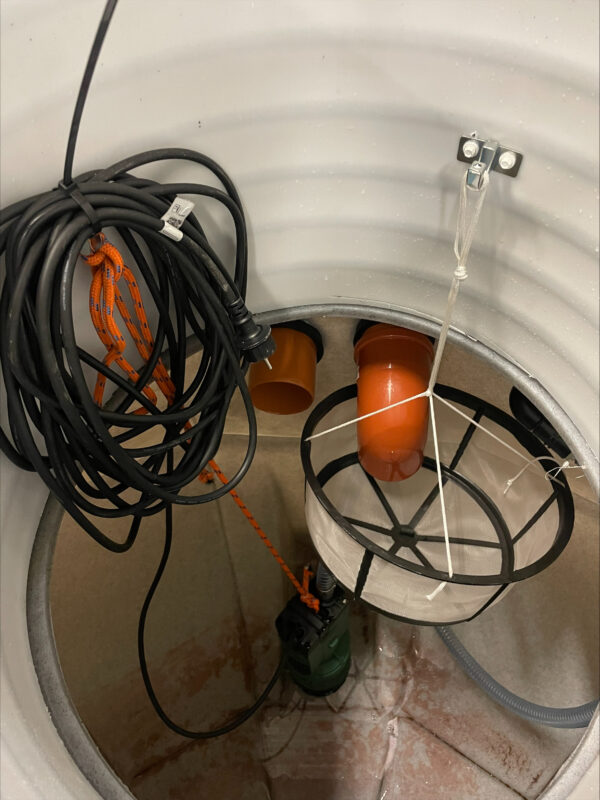 En närbild av ÄlvestadTanken regnvattentank CL 3500L lågbyggd interiör, som visar två orange rör, en svart kabel, en nätfilterkorg som hänger inuti och en grön dränkbar pump som är fäst vid ett orange rep.