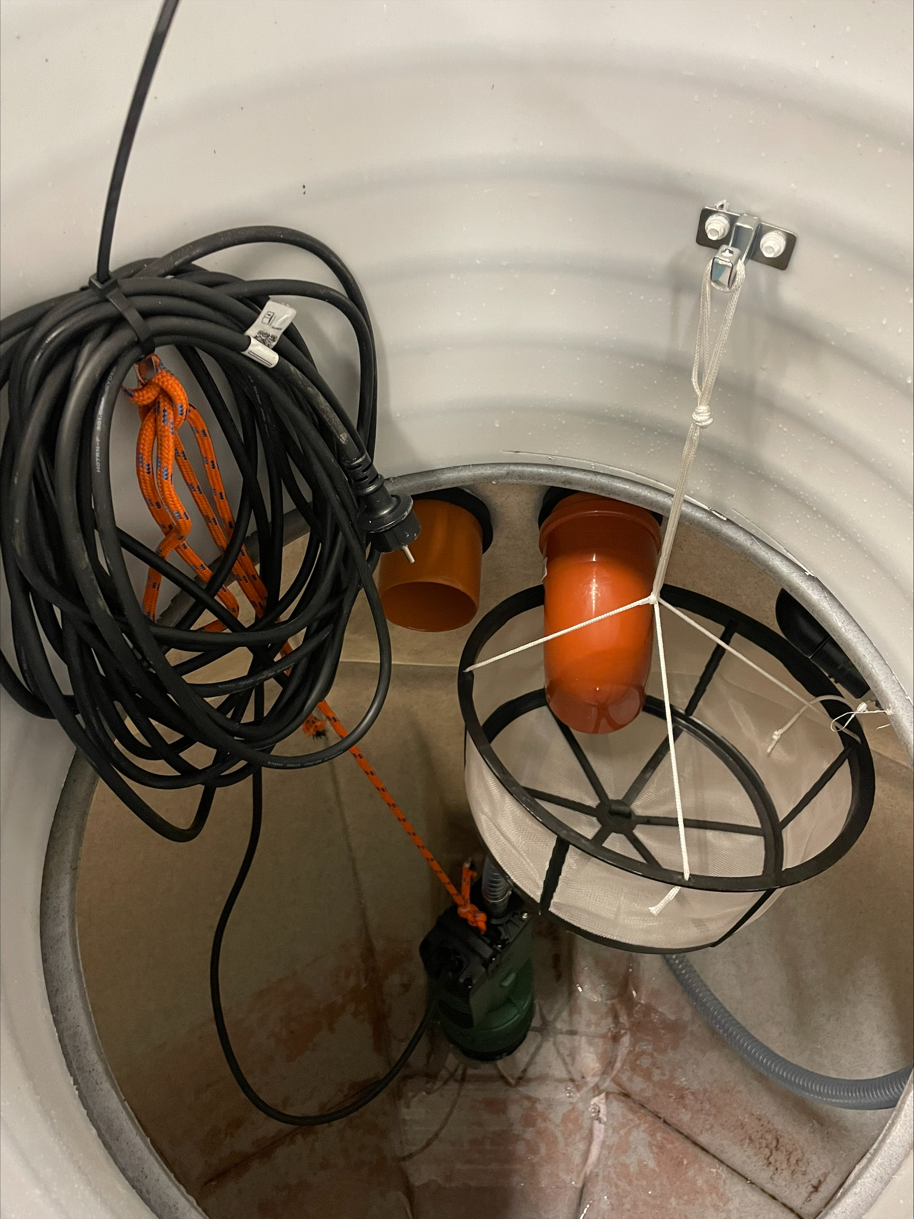 En närbild av ÄlvestadTanken regnvattentank CL 3500L lågbyggd interiör, som visar två orange rör, en svart kabel, en nätfilterkorg som hänger inuti och en grön dränkbar pump som är fäst vid ett orange rep.