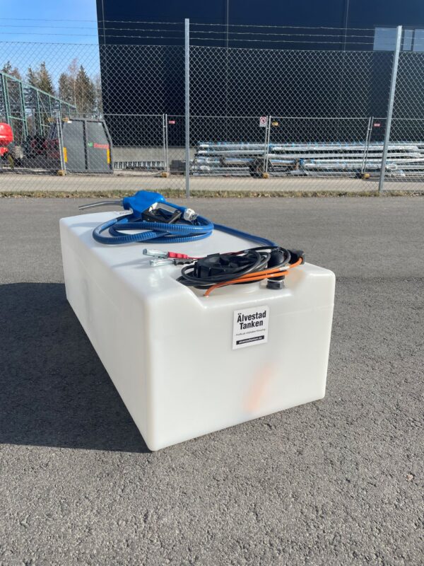 En stor rektangulär vit ÄlvestadTanken AdBluetank 200L Låg med slangar och ett blått munstycke på toppen är placerad på asfalt nära ett kedjestängsel och industribyggnader under klar himmel.
