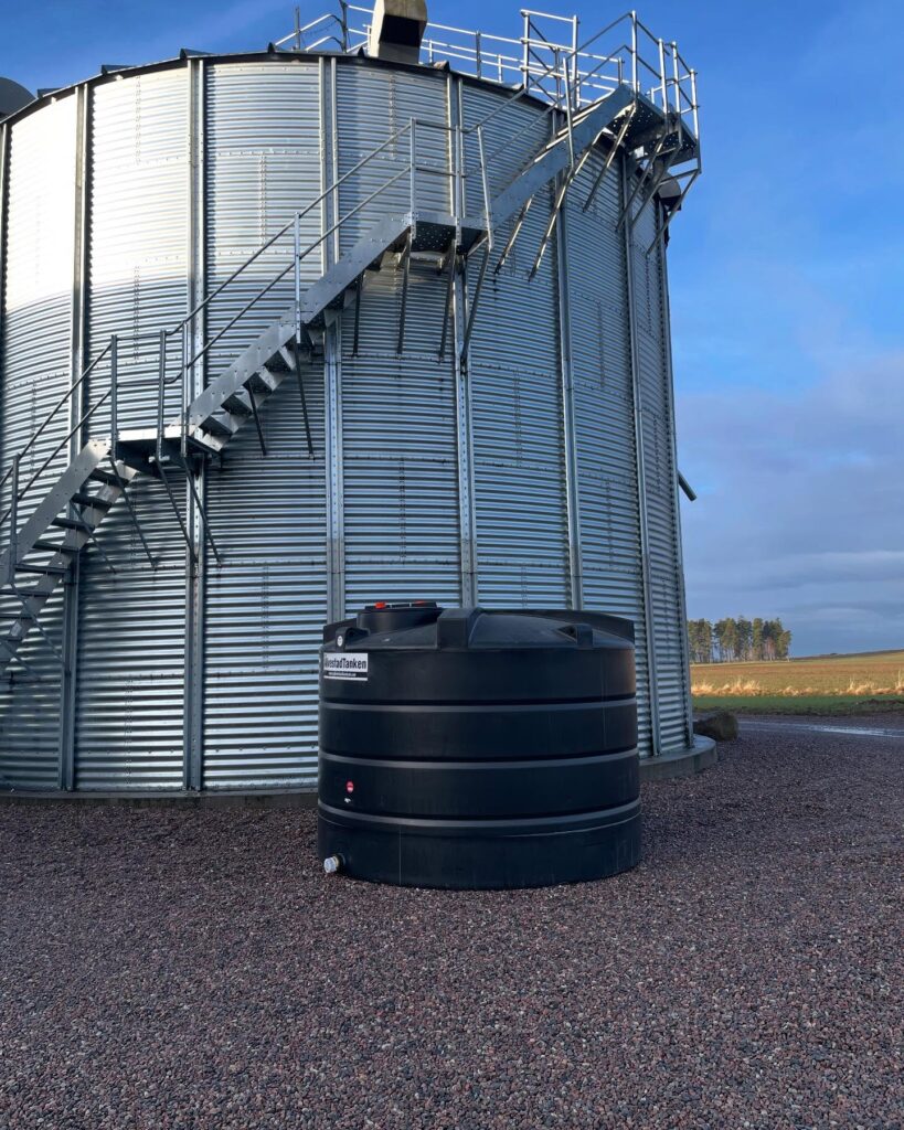En Bufferttank 7000 liter - komplett vattentank är placerad på grus bredvid en hög spannmålstank av korrugerad metall med utvändig trappa, allt under en blå himmel i ett lantligt landskap.