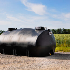 En stor svart Uthyrning - Vattentank TankMaster 6000L vattenlagringstank står på grus framför ett grönt fält under en blå himmel med lätta moln.