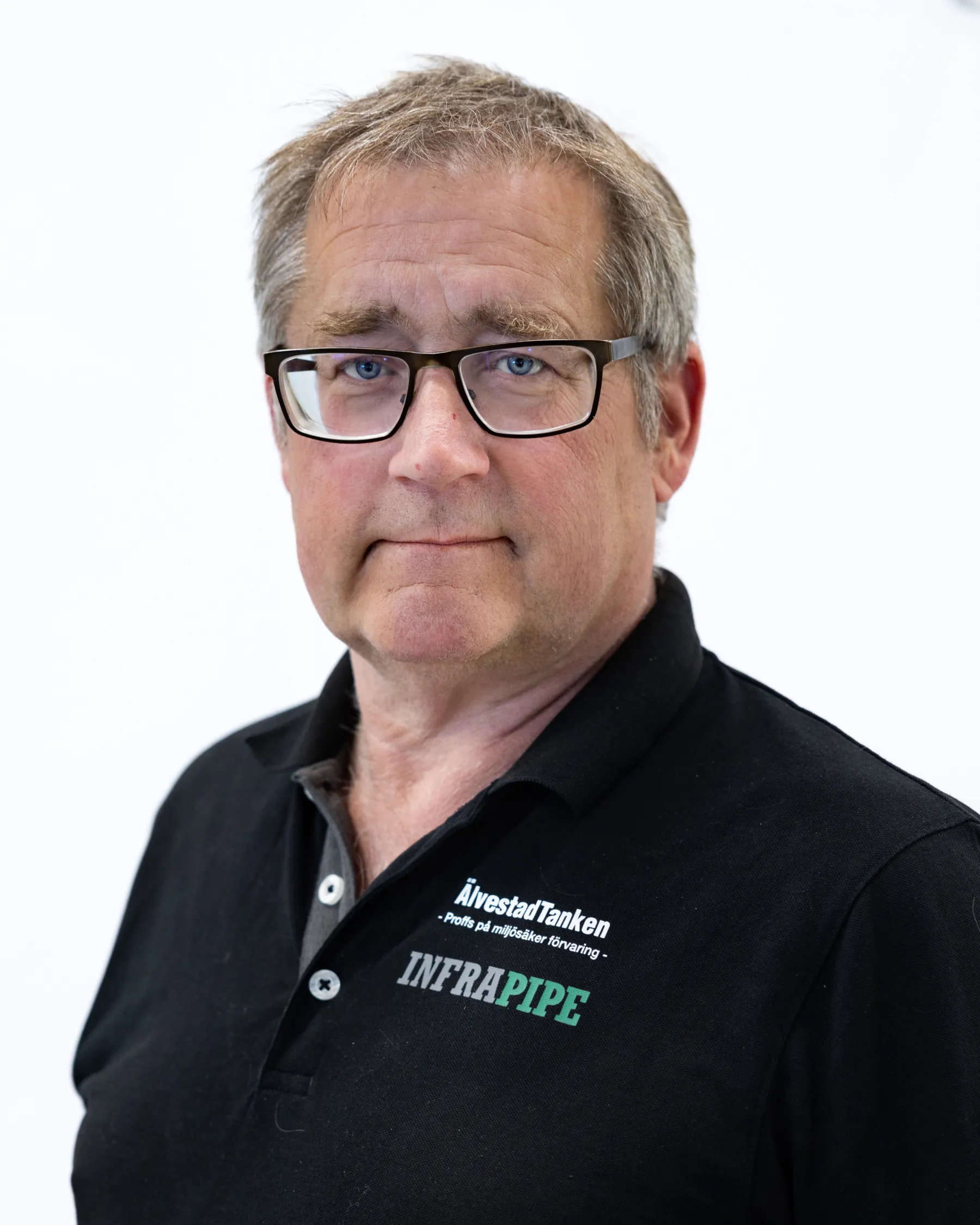 En medelålders man med kort grått hår och glasögon, iklädd en svart polotröja med logotyperna ÅlvestadTanken och INFRAPIPE, står mot en vit bakgrund.