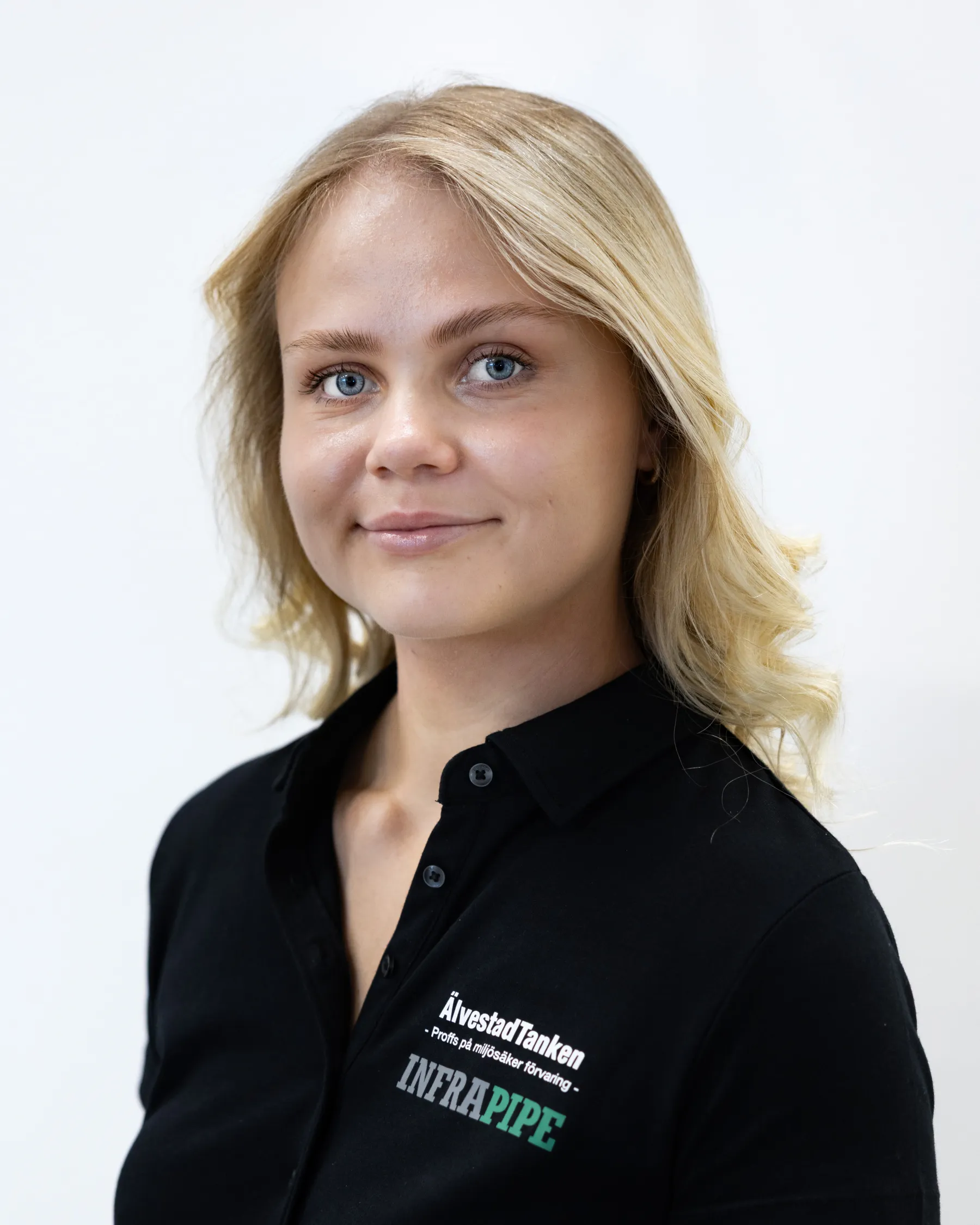 En ung kvinna med blont hår i lösa vågor, klädd i en svart skjorta med vit och grön logotyp med texten ÄlvestadTanken och INFRAPIPE, poserar mot en vit bakgrund.