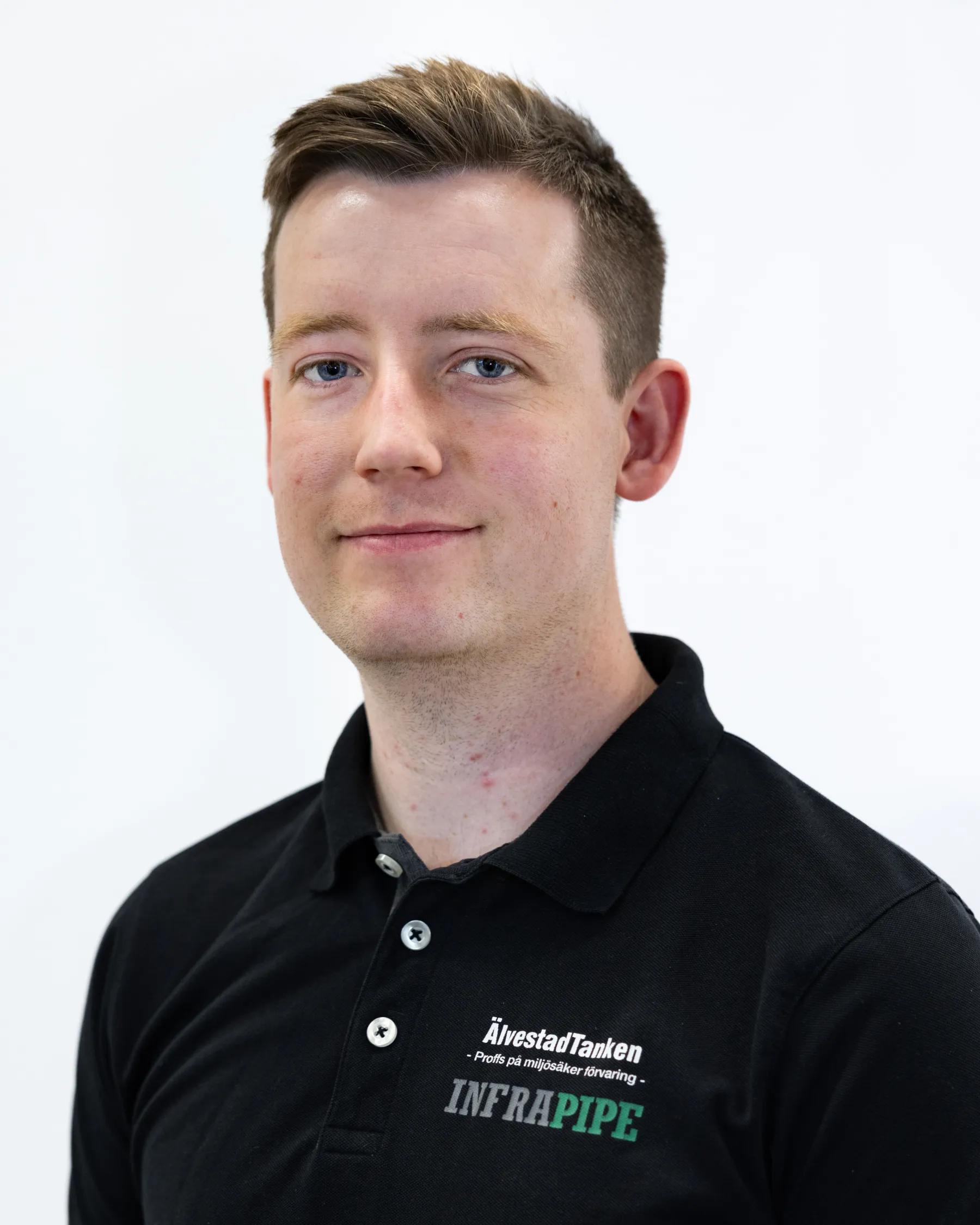 En ung man med kort brunt hår, klädd i en svart skjorta med krage och logotyperna ÄlvestadTanken och INFRAPIPE, står mot en vit bakgrund, vänd mot kameran och ler lätt.