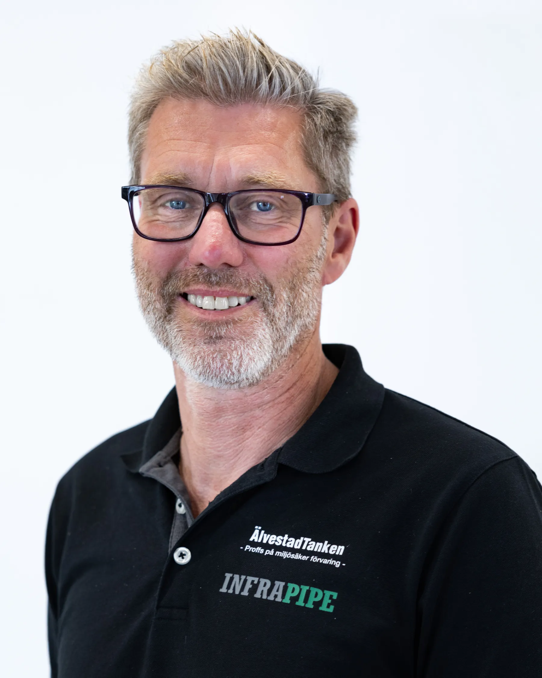 En medelålders man med grått hår, skägg och svarta glasögon ler mot kameran. Han bär en svart polotröja med vita och gröna logotyper där det står ÄlvestadTanken och INFRAPIPE. Bakgrunden är enfärgad vit.