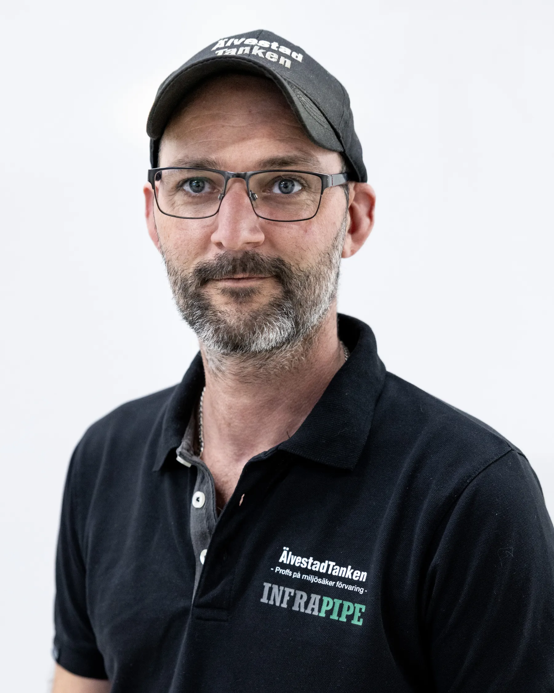 En man med skägg, glasögon, svart keps och svart polotröja med ÄlvestadTankens och INFRAPIPE:s logotyper, står mot en vit bakgrund.