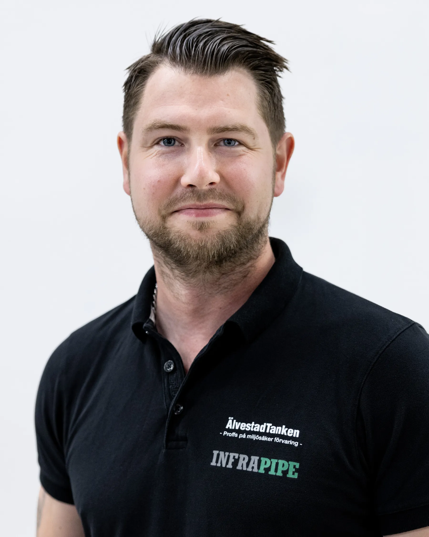 En man med kort, stylat brunt hår och skägg, klädd i svart polotröja med logotyperna ÄlvestadTanken och INFRAPIPE, poserar framför en vit bakgrund.