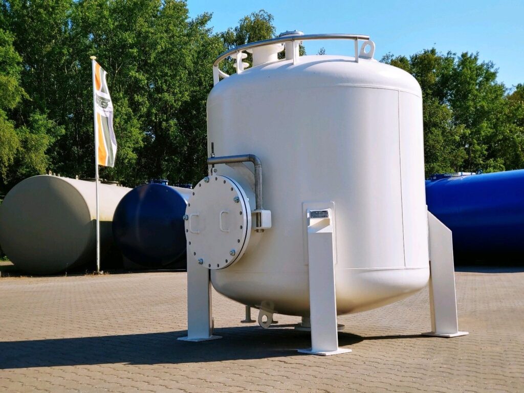 En stor, vit, cylindrisk industritank med en bultad tillträdeslucka står på en asfalterad yta utomhus. I bakgrunden syns andra cisterner och en flagga, gröna träd och en klarblå himmel.