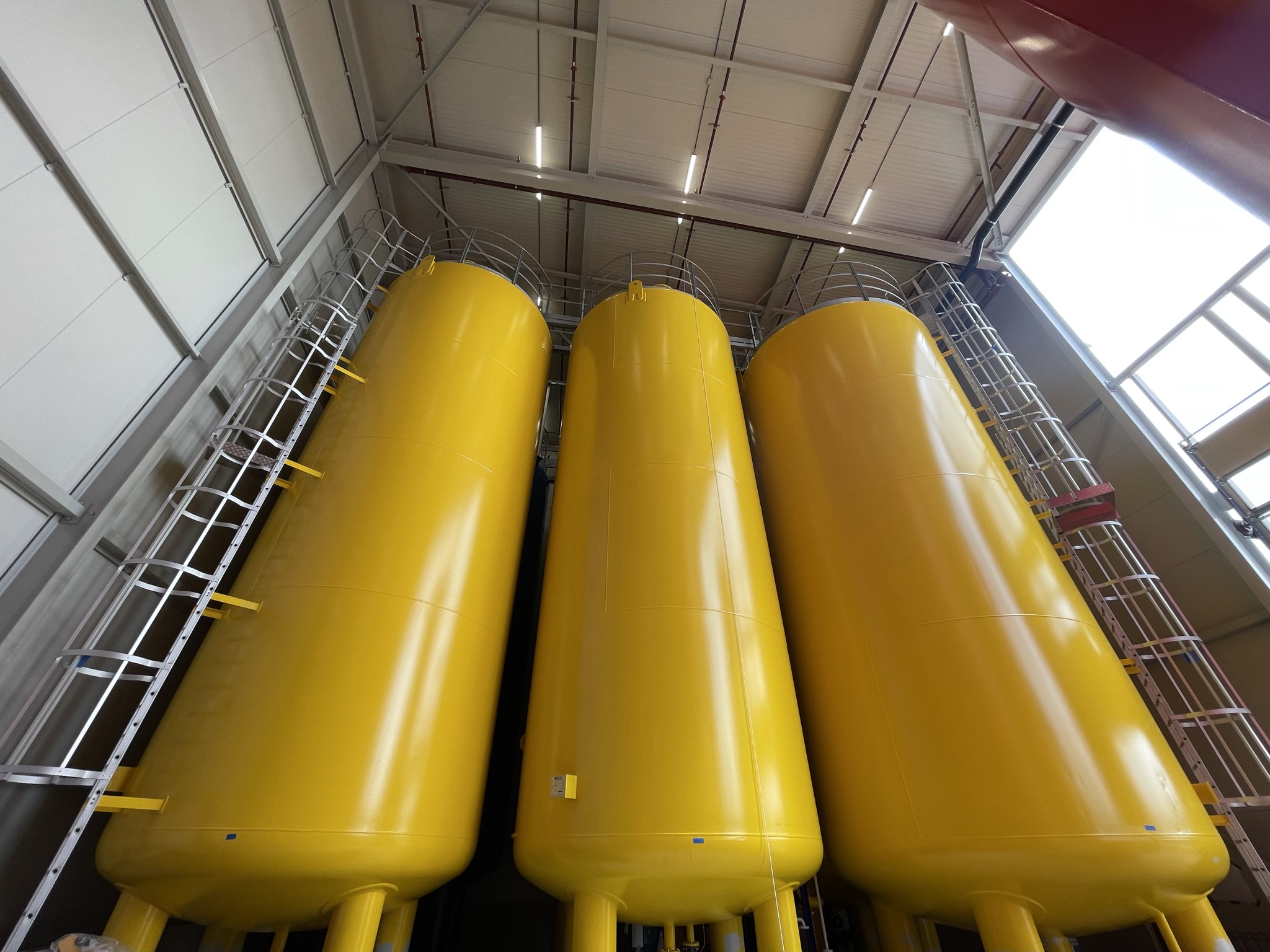 Tre stora, ljusgula industriella lagringstankar står sida vid sida inomhus, med metallstegar och räcken längs sidorna, mot ett högt tak med taklampor.