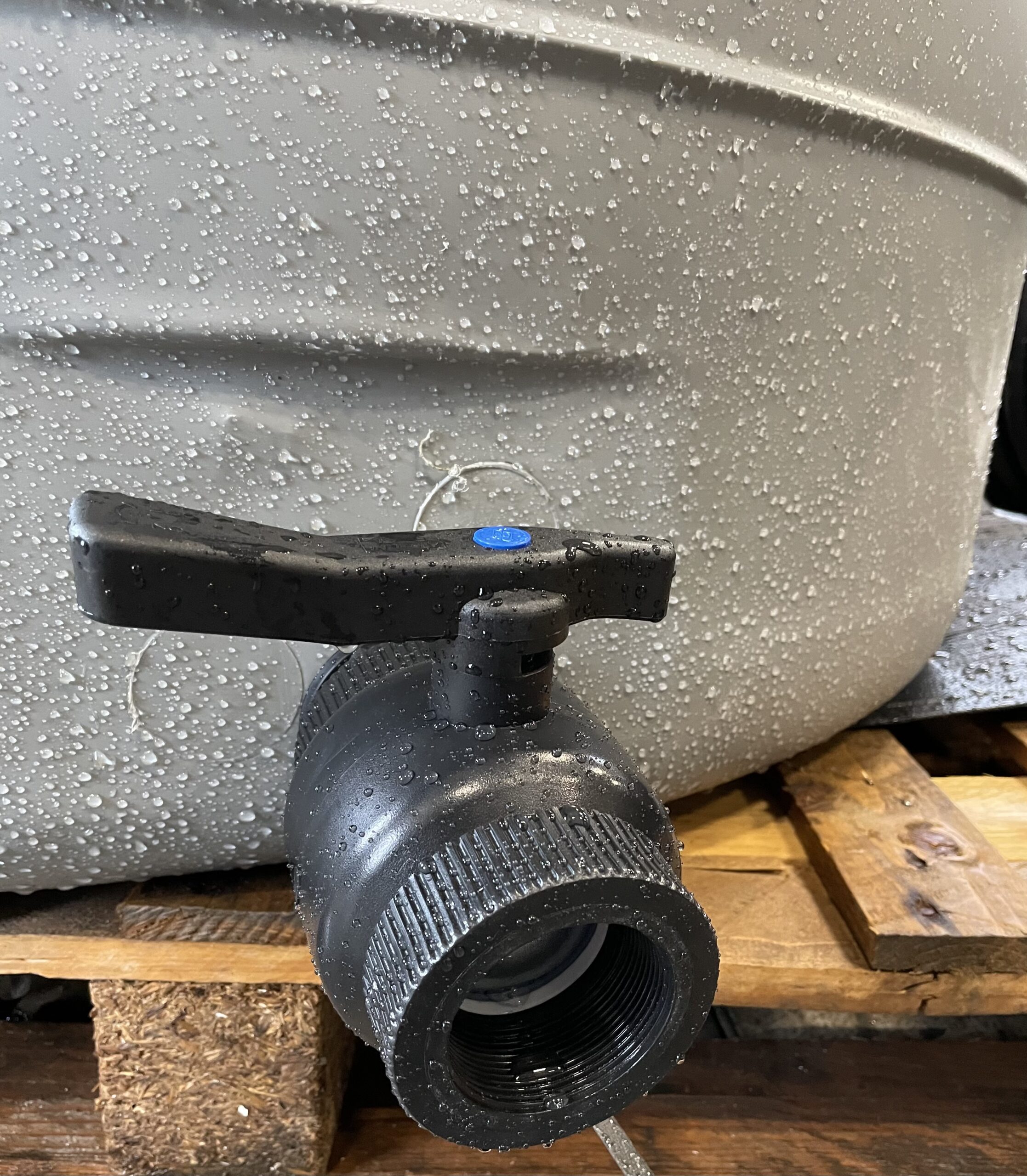 En närbild av Vattentank V 2000L - Visningsexemplar visar en svart plastventil på en grå, vattenspräcklig behållare placerad på en träpall. Ventilhandtaget är horisontellt och det finns vattendroppar på både ventilens och tankens yta.