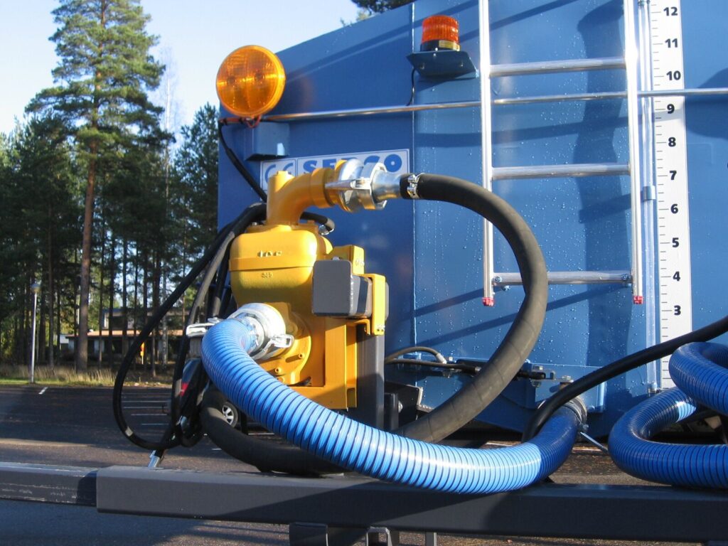 En närbild av JCE pumpens hydraldrivna slangar och en gul pump ansluten till en blå tank med stege och mätare, utomhus med träd och klar himmel i bakgrunden.