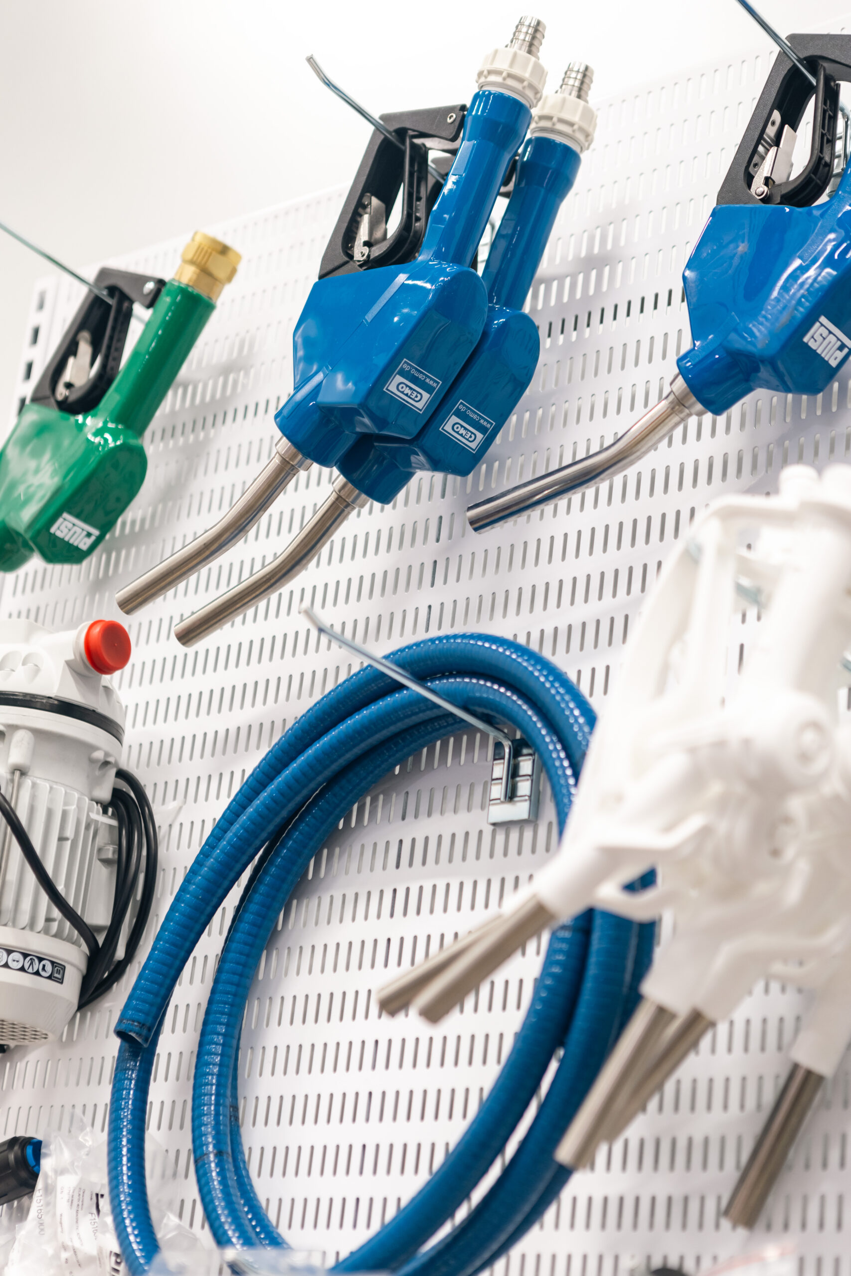 Flera blå och gröna bränslemunstycken och upprullade blå slangar visas på en perforerad vit vägg, tillsammans med mekaniska komponenter, i en verkstads- eller butiksmiljö.