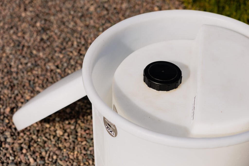 Närbild av "Säkerhetsinvallning för doseringstank", en vit plastbehållare med svart skruvlock och handtag, som står på grus utomhus i solljus. Behållaren ser något sliten ut.