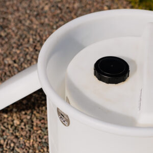 Närbild av "Säkerhetsinvallning för doseringstank", en vit plastbehållare med svart skruvlock och handtag, som står på grus utomhus i solljus. Behållaren ser något sliten ut.